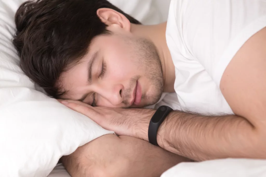 Handsome young guy sleeping in comfortable bed, man lying on cozy pillow with eyes closed, wearing white t-shirt and smart wristband tracker for sleep tracking, keeping healthy living, day regimen